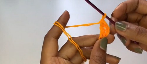 Hands working orange yarn around a crochet hook to form the beginning chain on a white surface