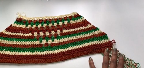 Flat lay of a crochet fringe top panel in progress showing rust brown green and cream stripe and granny stitch sections with fringe strands hanging from the bottom edge