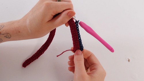 Hands holding a pink crochet hook working a two-color ribbed strip in burgundy and navy yarn