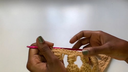 Hands using a pink crochet hook to work across a row of open granny stitch in golden yellow yarn