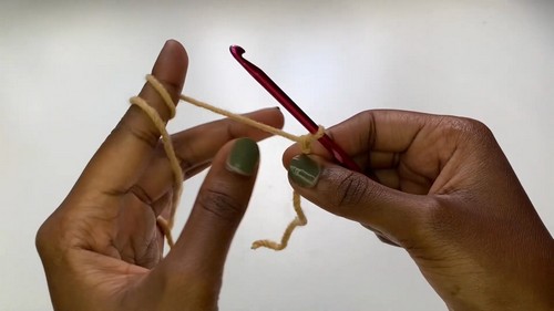 Hands holding a red crochet hook working the first chain with natural beige yarn against a white background
