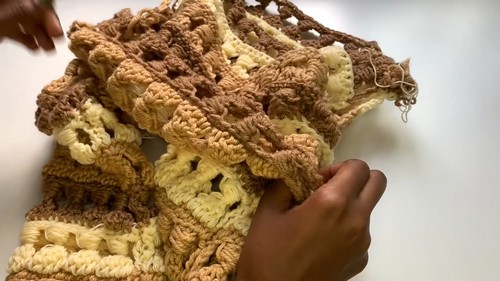 Hands holding a partially assembled crochet cold shoulder top in brown tan and cream granny stitch sections with loose yarn ends visible