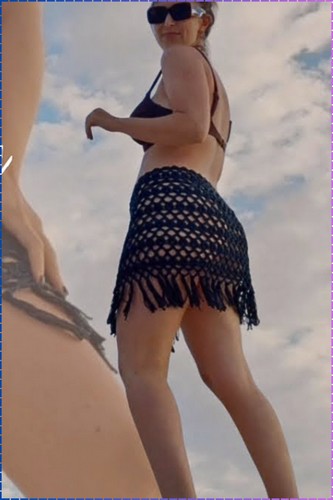 Woman wearing a black crochet mesh mini skirt with fringe hem over a bikini bottom photographed from below against a cloudy sky at the beach