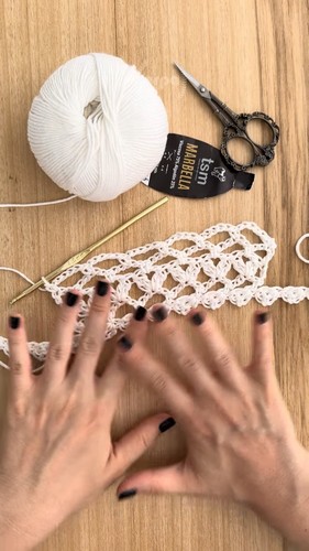 Hands smoothing out a completed white crochet lace strip beside a ball of Marrella yarn and scissors on a wooden surface