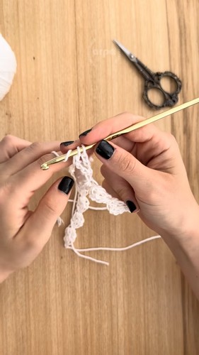 Hands using a gold crochet hook to work a delicate lace stitch pattern in white yarn with scissors visible in the background on a wooden surface