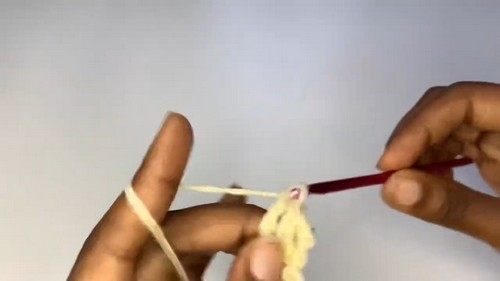 Hands holding a red crochet hook working the first chain stitches with pale yellow yarn against a light background