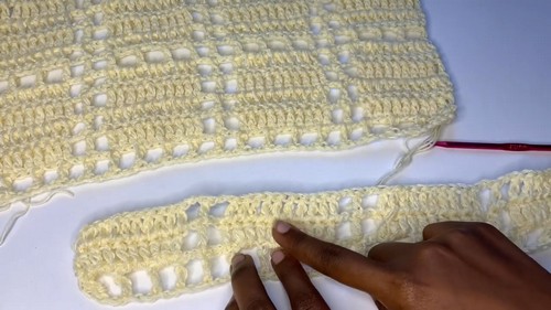 Hands holding a completed pale yellow crochet panel with open mesh stitch pattern next to a smaller strap piece with a red hook visible at the edge